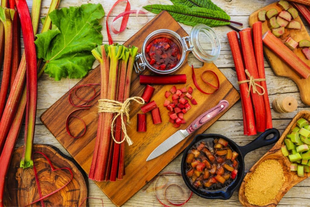 egaturi de rubarba pe tocator si masa din lemn, alaturi de borcan cu dulceata de rubarba